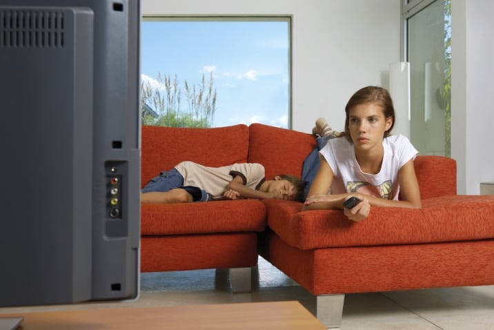 a girl watching TV while her brother is sleeping on the sofa